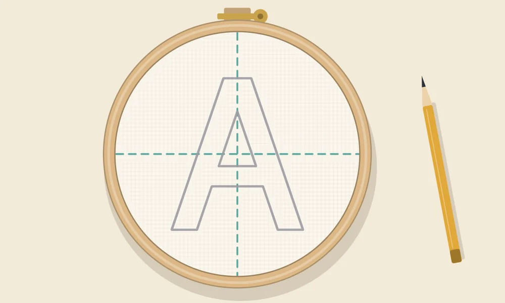 Letter A traced in pencil on hooped fabric, centered on a vertical and a horizontal dashed guide line that cross in the middle