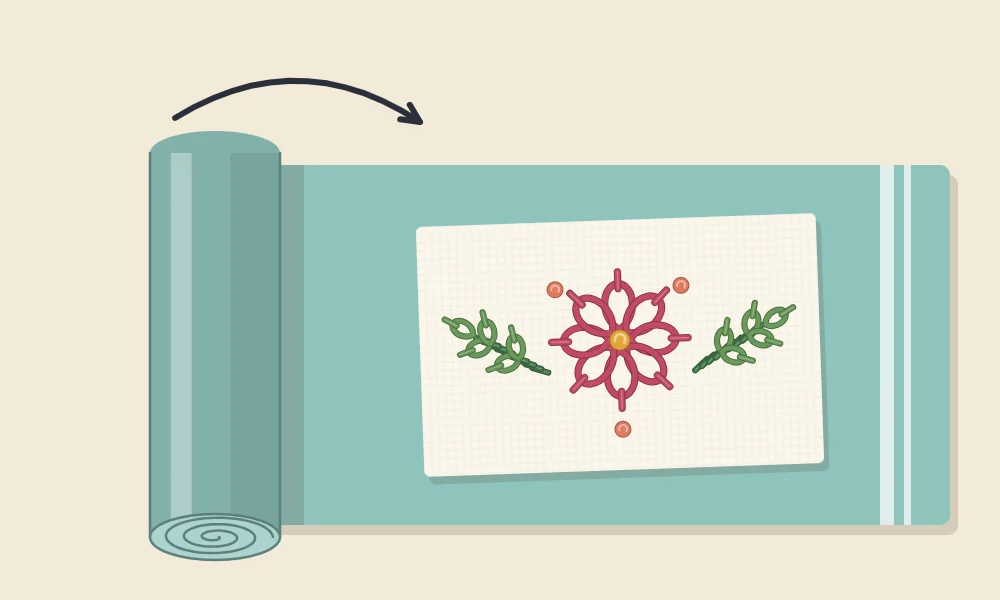 Damp embroidery laid on a towel that is being rolled up from one end, with a curved arrow showing the rolling direction